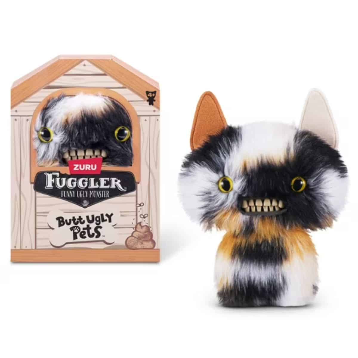 A furry calico Fuggler toy with mismatched ears, yellow eyes, and a wide toothy grin shown beside its packaging from the Butt Ugly Pets collection.