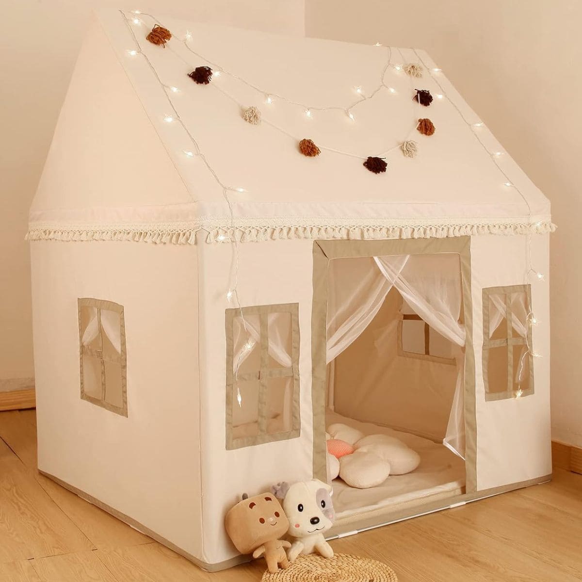 A beige kids play tent shaped like a little house with fabric windows, soft floor cushions, string lights, and stuffed toys placed at the front.