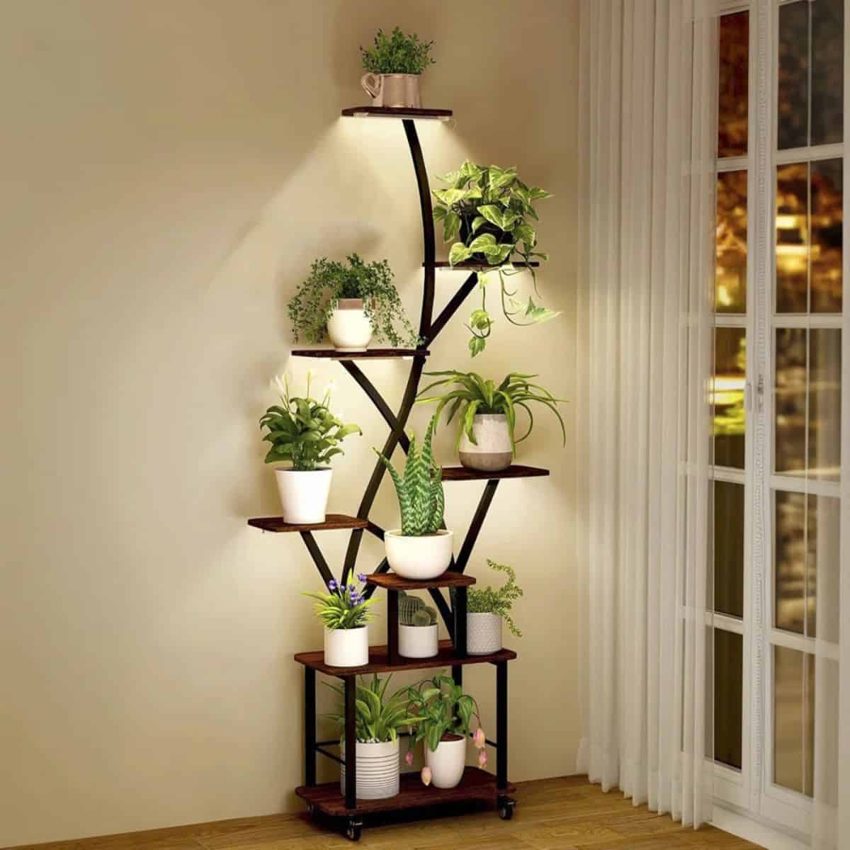 A tall corner plant stand with built-in lights, holding multiple potted houseplants on tiered wooden shelves, set beside a window with sheer curtains.