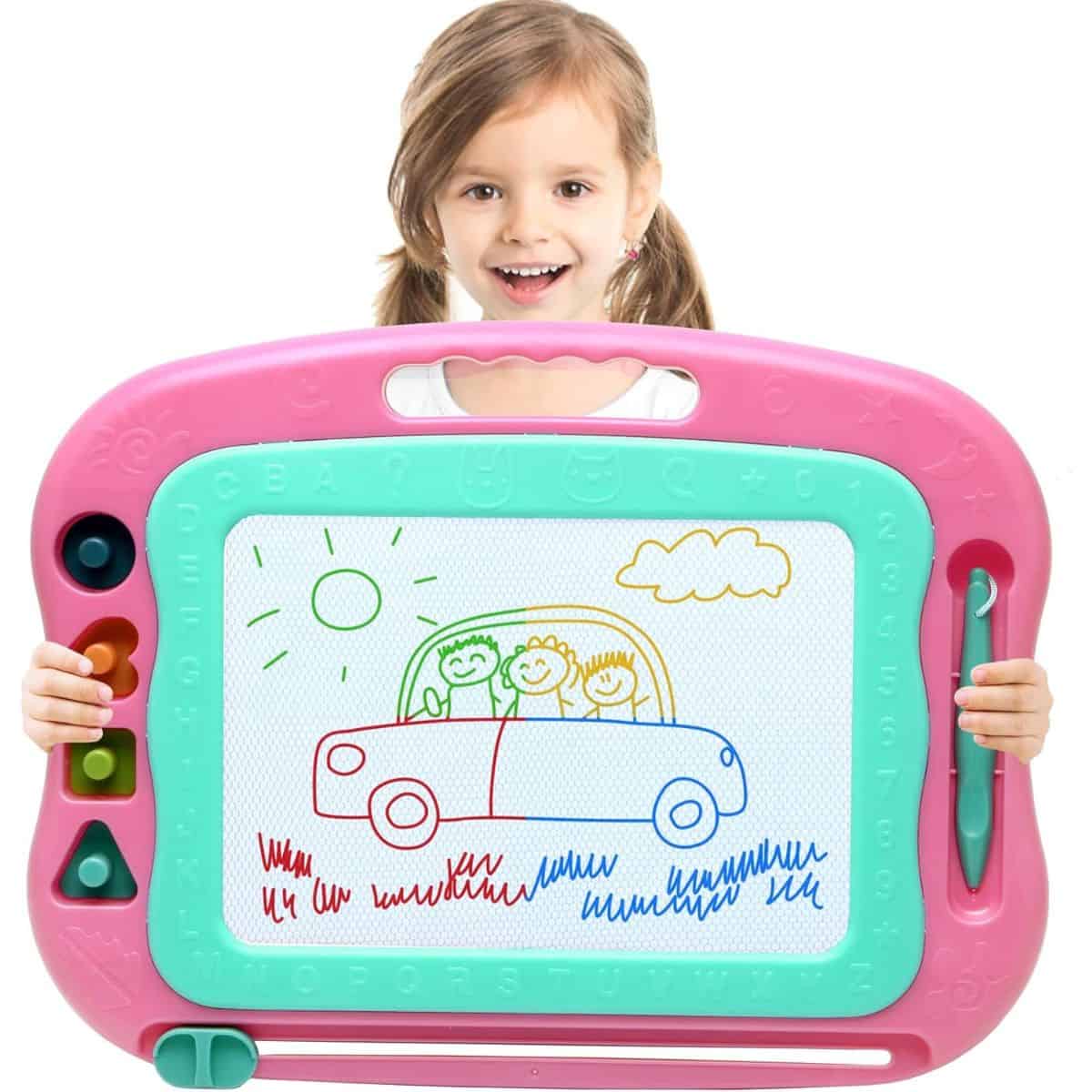 Young girl holding a large pink and teal magnetic drawing board featuring a colorful doodle of a family in a car under the sun and a cloud.