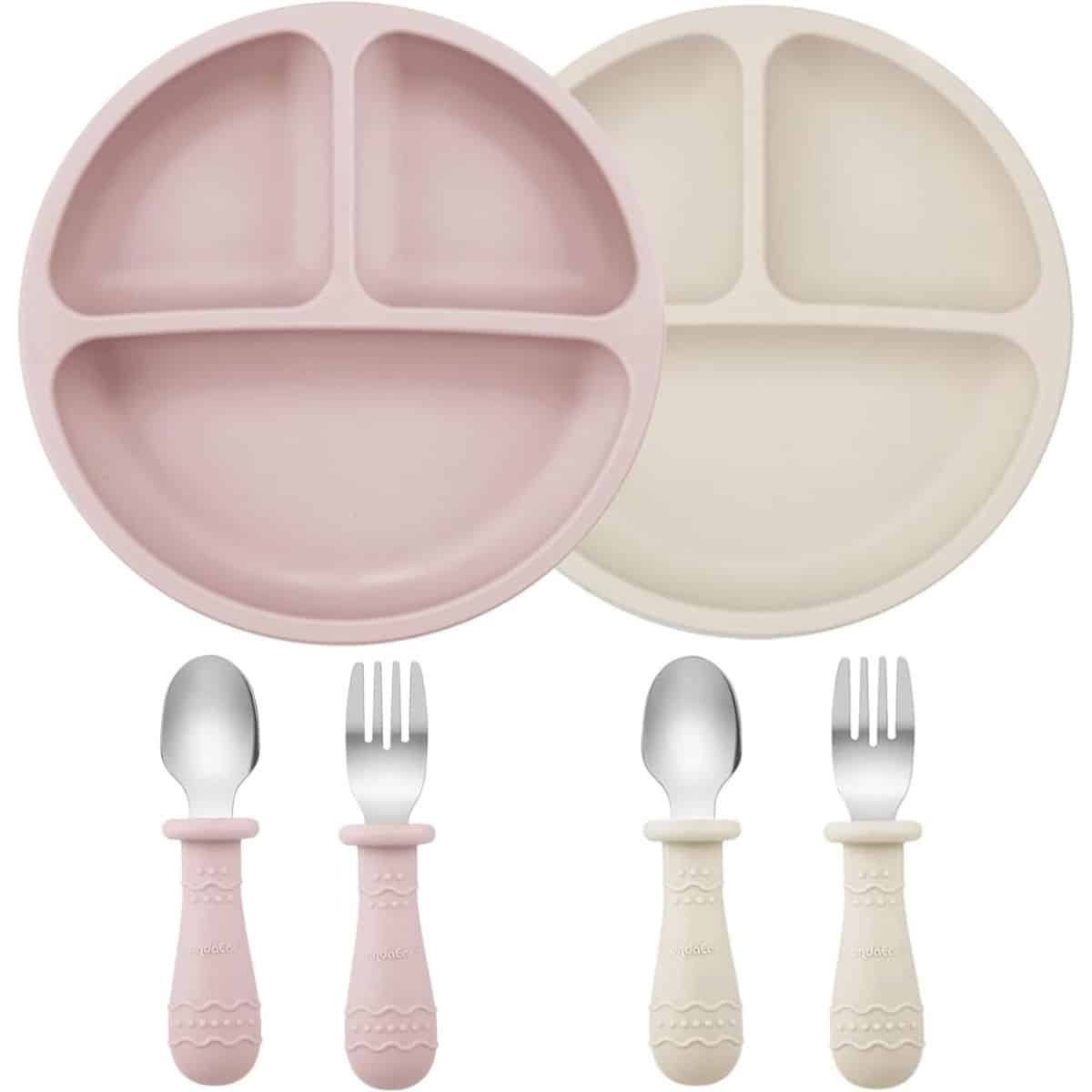 Two toddler divided plates in pink and cream with matching short-handled spoons and forks, all designed for easy gripping and safe self-feeding.
