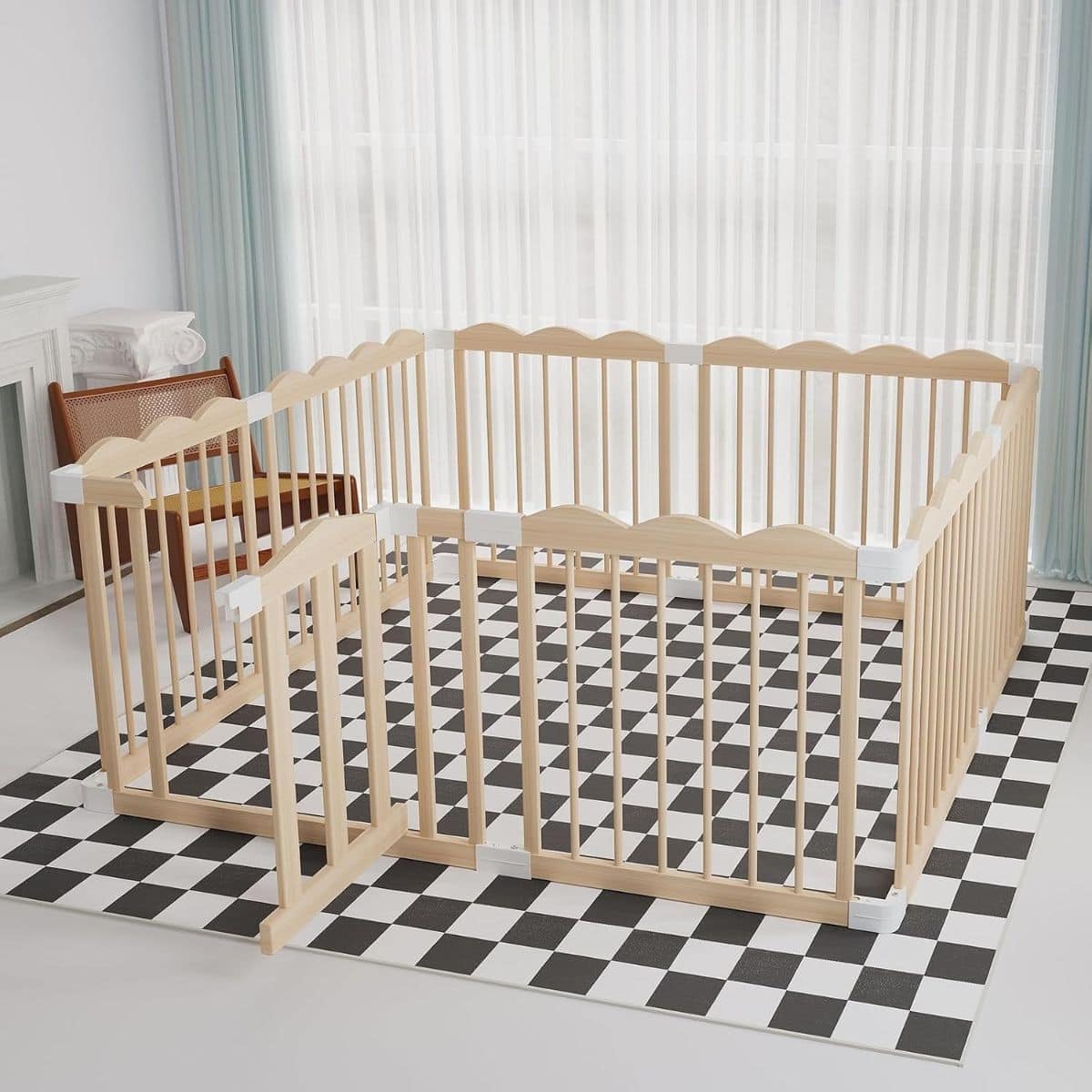 Natural wood baby playpen with an open gate and white corner connectors on a black and white checkered floor mat.