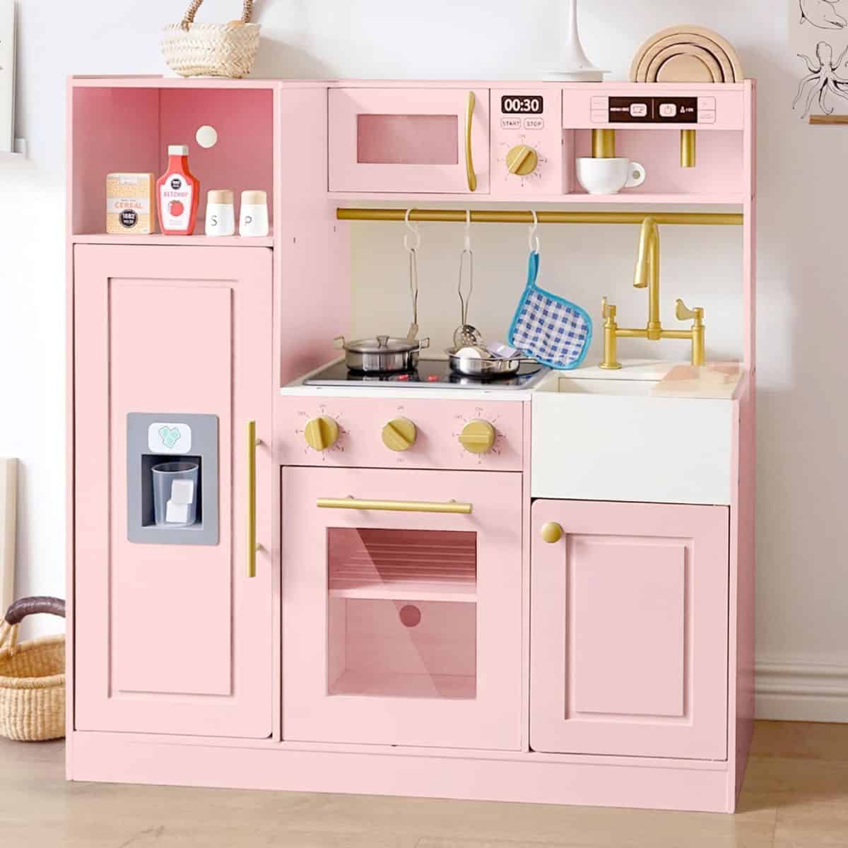 A pink wooden play kitchen with a stove, oven, sink, microwave, fridge, and toy accessories like pots, dishes, and a potholder arranged neatly.
