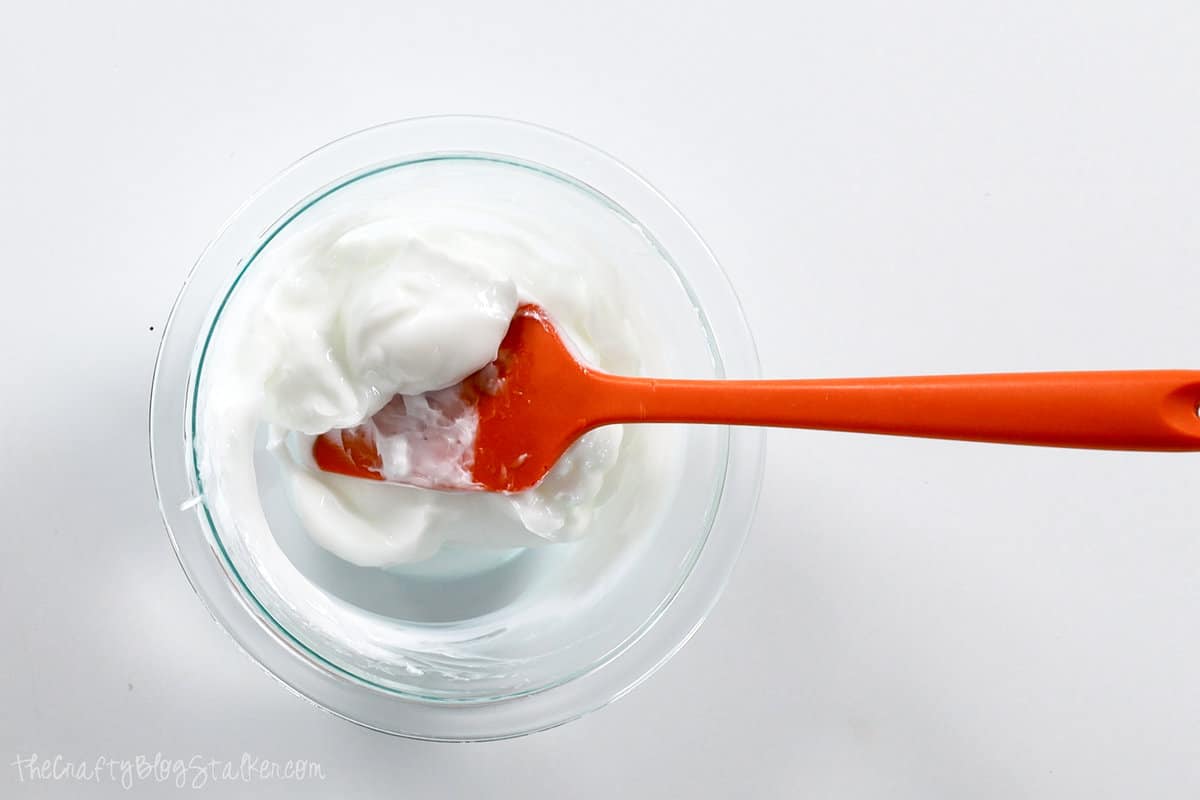 A glass bowl with lotion and an orange spatula.