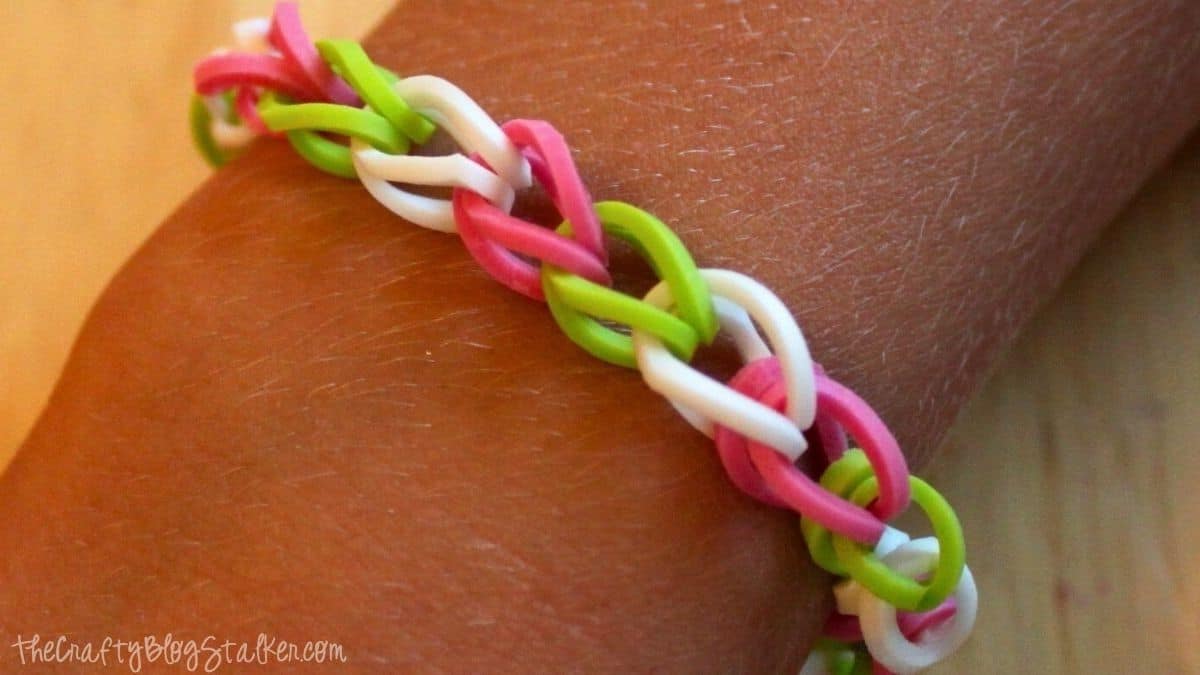 A close-up of a colorful loom band bracelet made with pink, white, and green rubber bands, worn on a wrist.