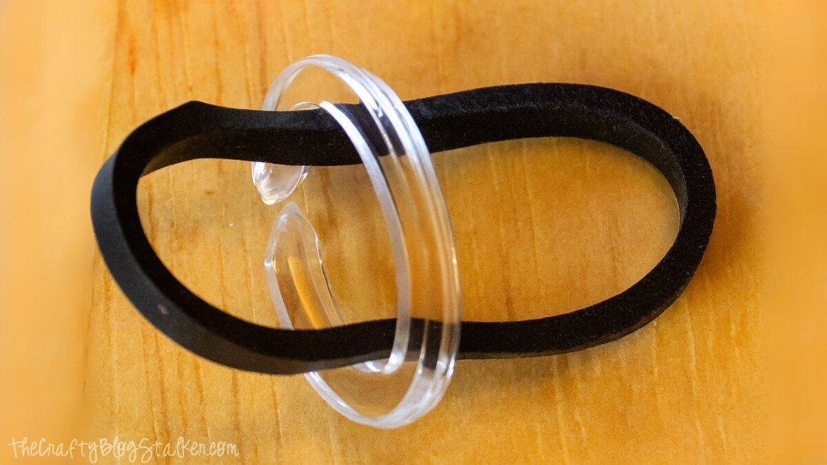 Close-up of a clear plastic C-clip holding a black rubber band, used for securing loom band bracelets on a wooden surface.
