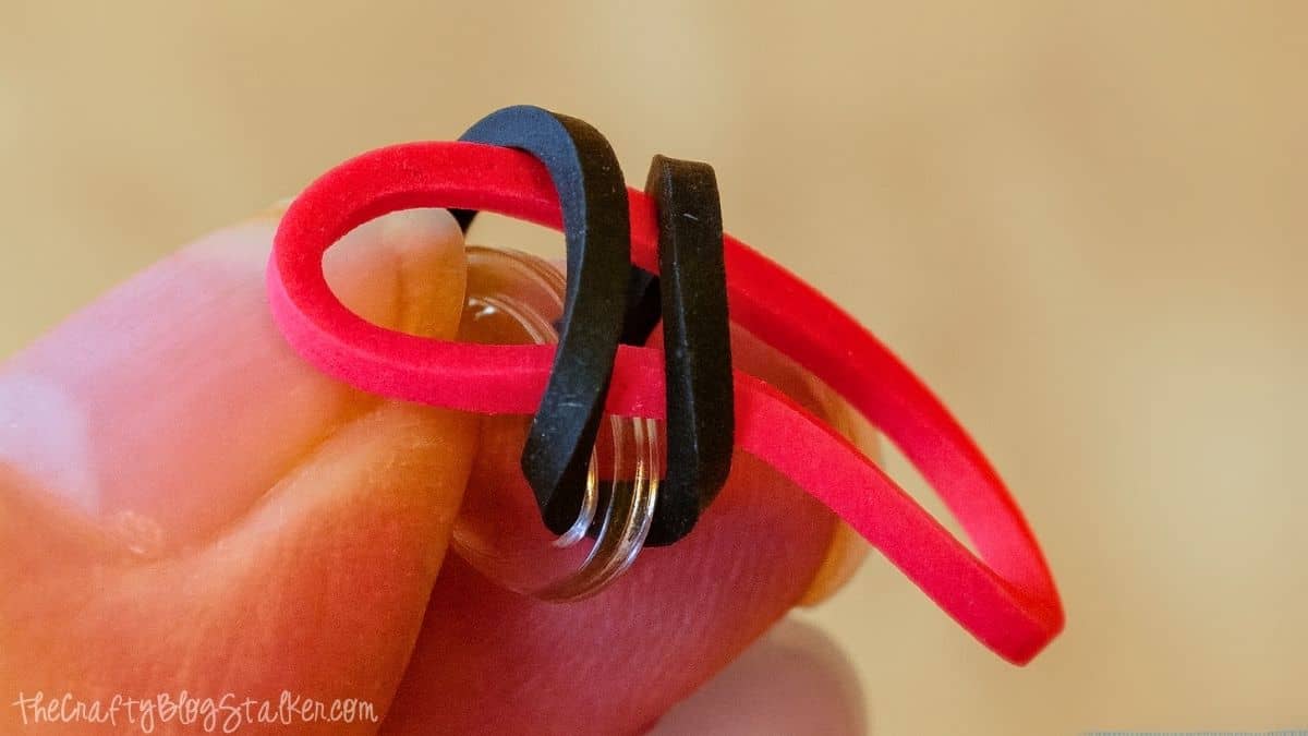 Close-up of fingers holding a clear plastic C-clip with a red and black rubber band looped through, showing the start of a loom band bracelet.