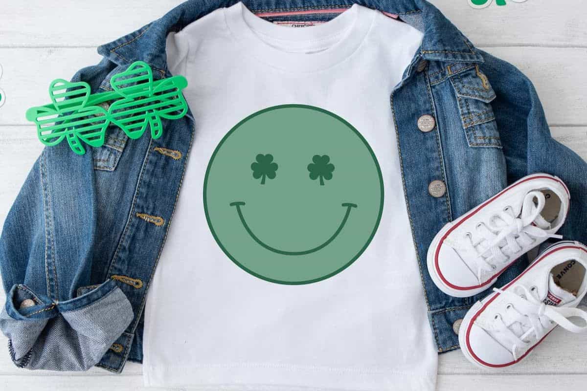 A white shirt with a design of a green smily face with clovers for eyes.