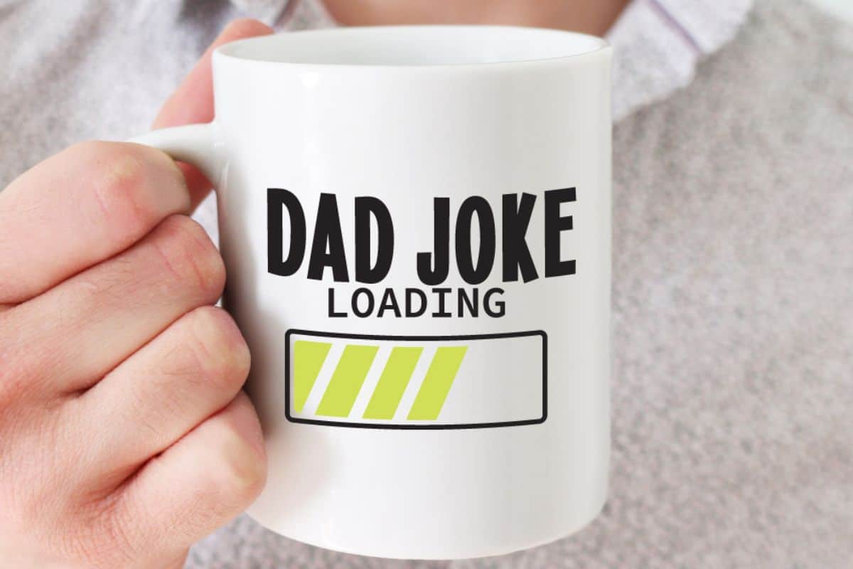 A hand holding a coffee mug that reads "Dad Joke Loading".