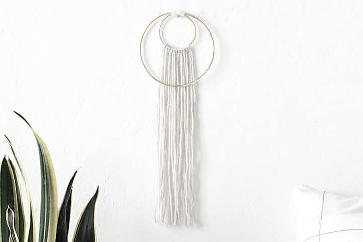 Minimalist double hoop wall hanging with white yarn strands, displayed on a white wall above a modern bed with a grid pillow and beside a tall snake plant.