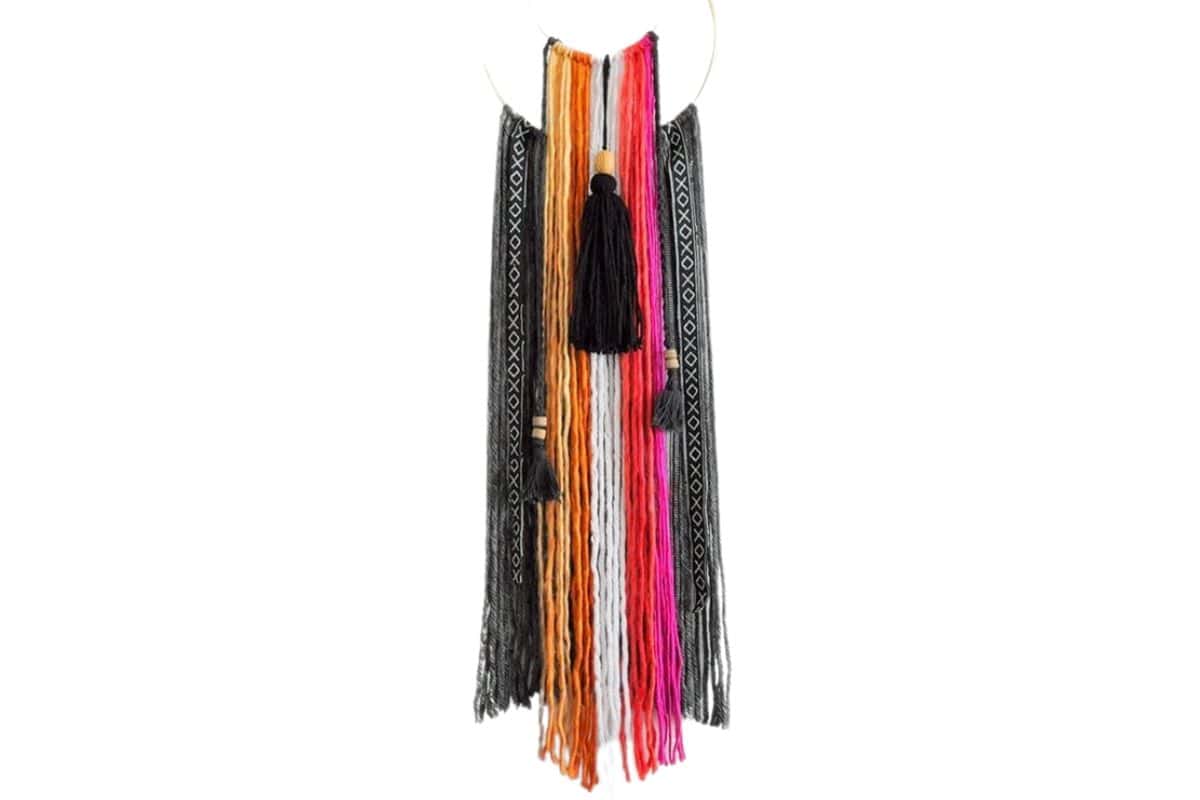 Colorful boho wall hanging with black, orange, pink, and white yarn strands, accented with tassels and patterned ribbon on a circular gold hoop.