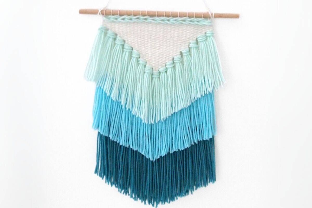 Layered tassel wall hanging made with shades of mint and teal yarn on a wooden dowel, forming a V-shaped design against a white background.