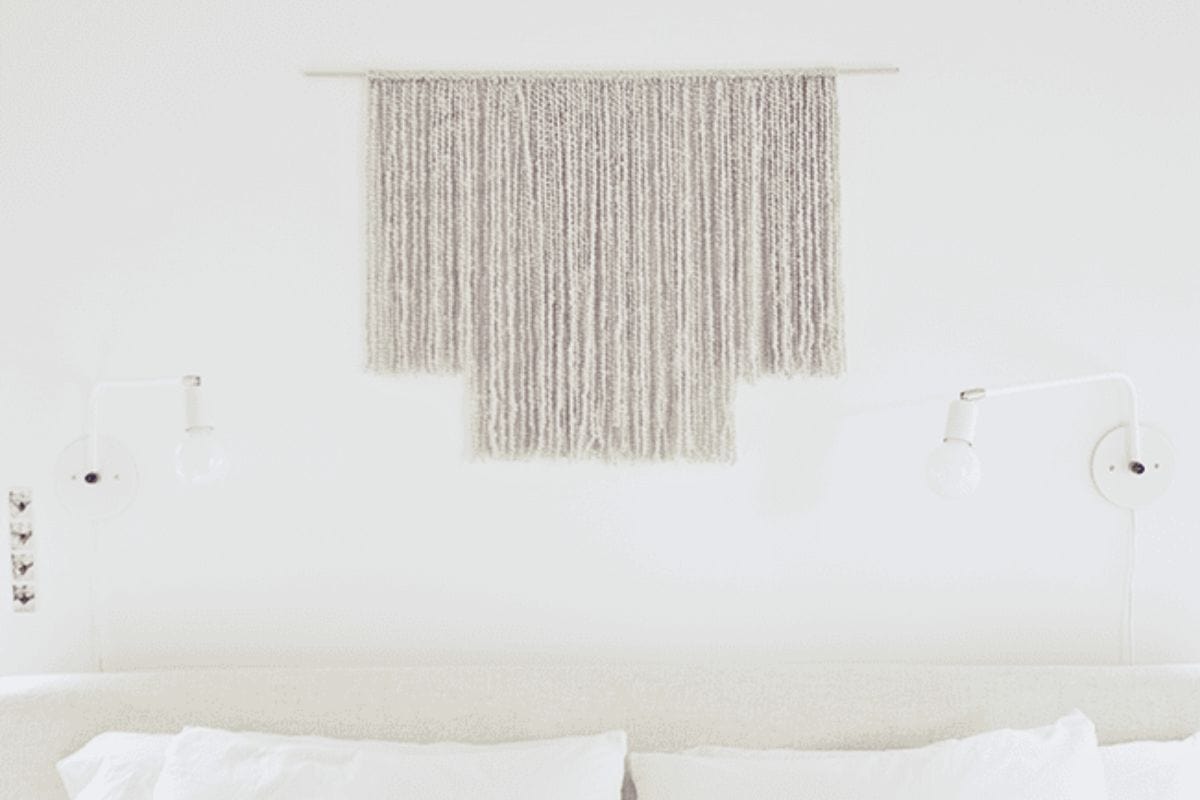 Minimalist gray woven yarn wall hanging displayed above a white bed with matching pillows and modern white wall-mounted reading lamps.