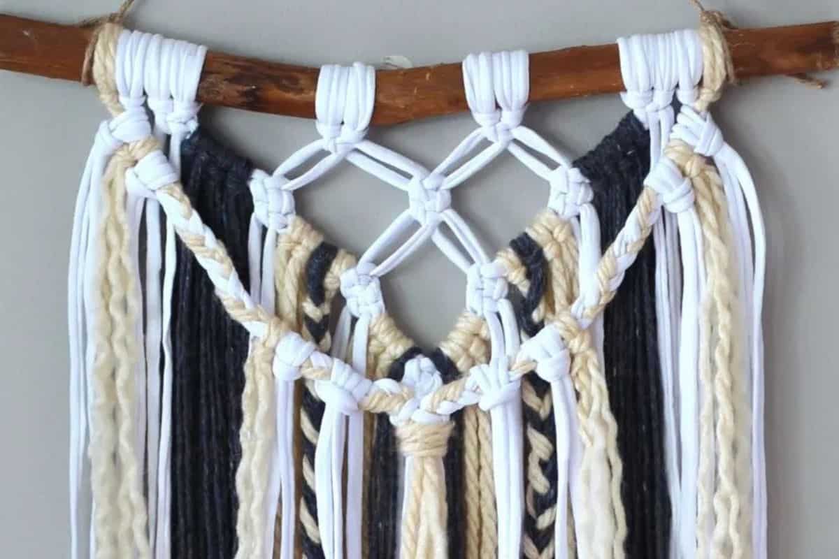 Close-up of a handmade macrame wall hanging with white, black, and beige yarn knotted onto a rustic wooden branch, featuring decorative diamond-shaped patterns.
