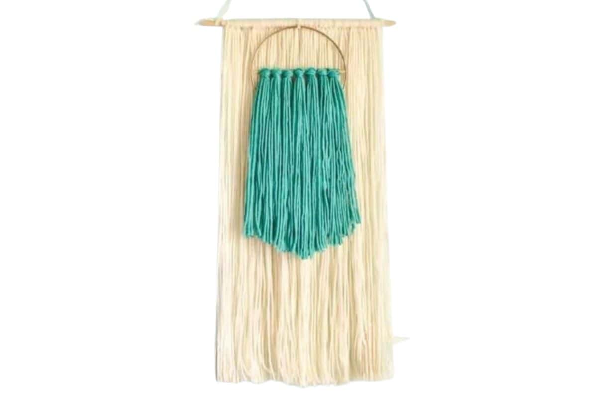 Boho-style yarn wall hanging with a layer of long cream strands and a smaller teal yarn design inside a gold ring, hanging from a wooden dowel.
