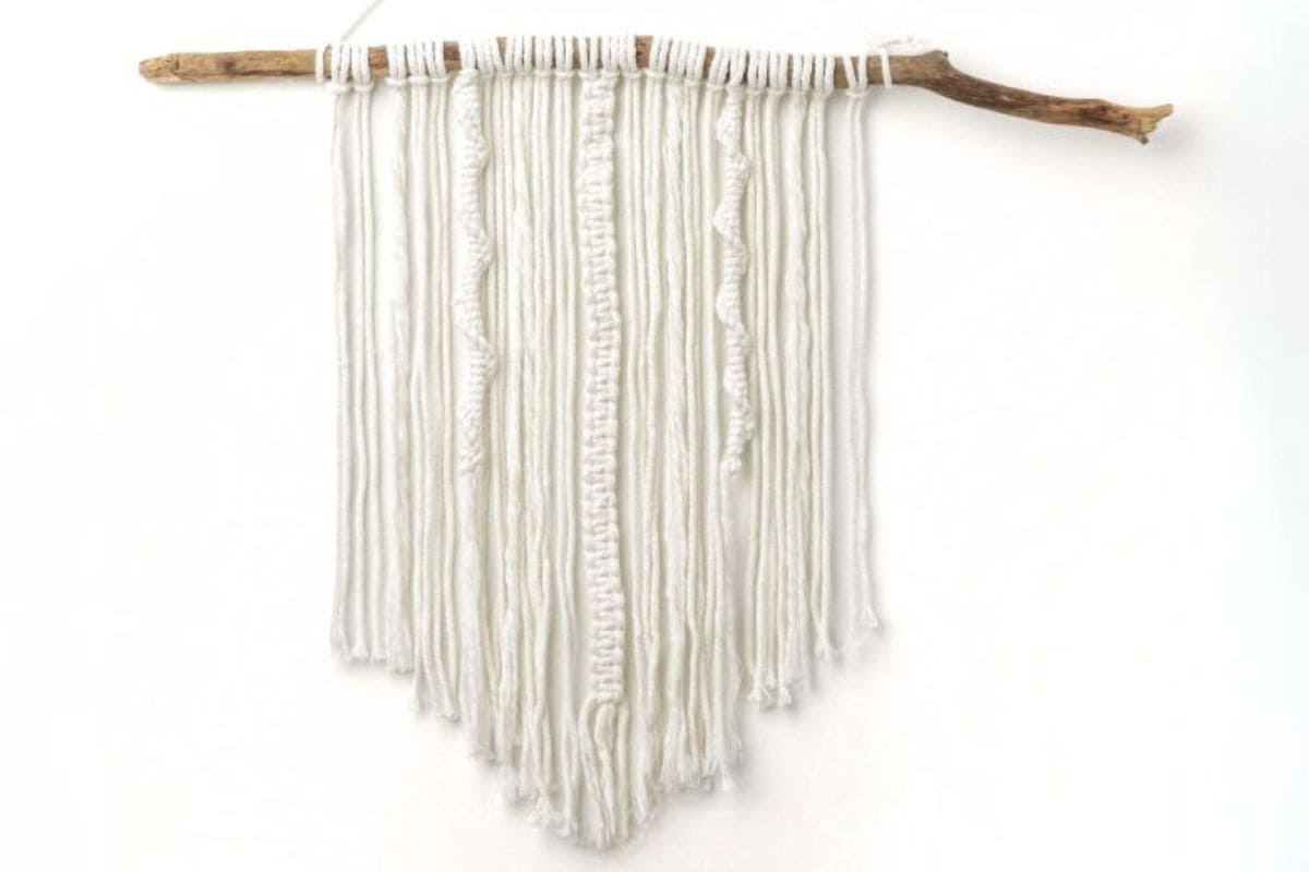 Minimalist macrame wall hanging made with white yarn strands and textured knots, suspended from a natural wooden branch against a white background.
