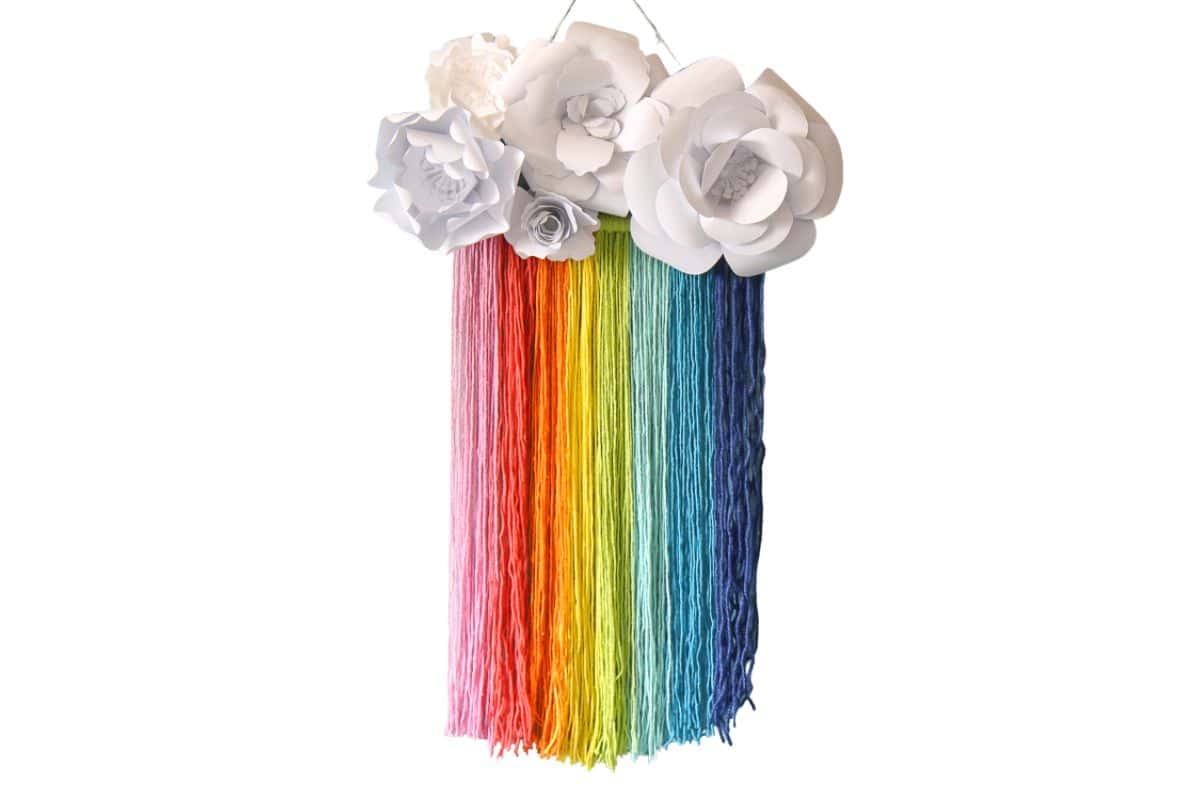 Colorful rainbow yarn wall hanging with strands in gradient hues from pink to blue, topped with large white paper flowers for a bright and whimsical decor accent.
