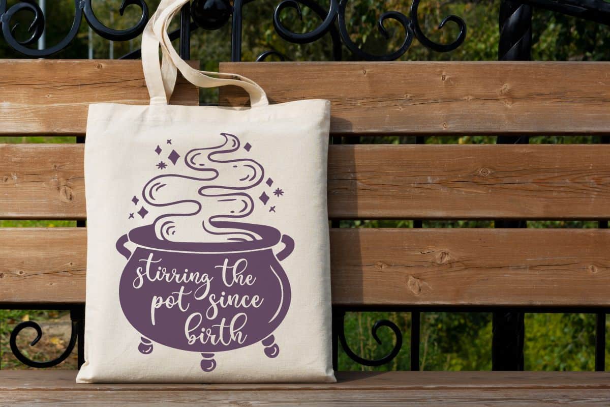 A tote bag on a park bench with the design - stirring the pot since birth.
