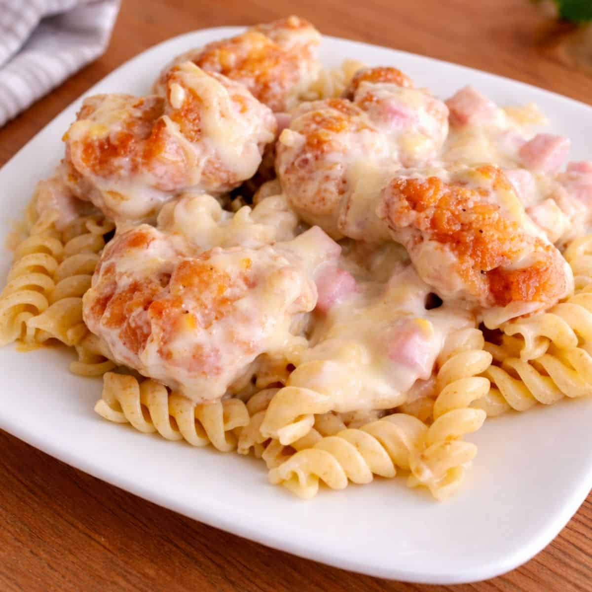 A plated serving of chicken cordon bleu casserole with crispy popcorn chicken, diced ham, melted cheese sauce, and rotini pasta on a white plate.