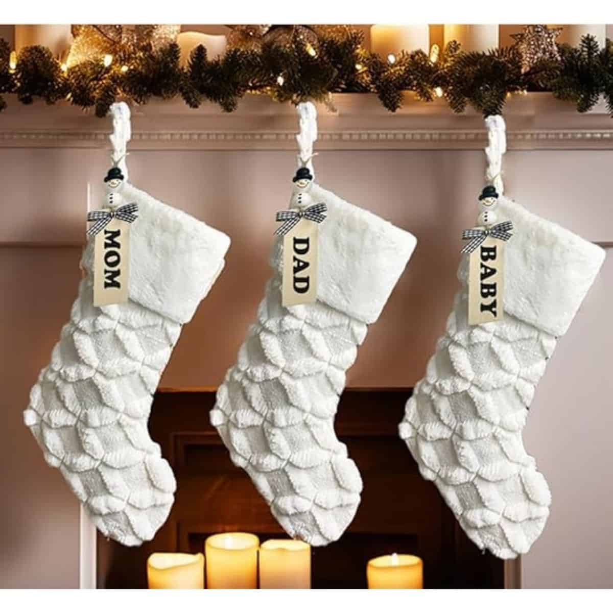 Three white faux fur Christmas stockings labeled Mom, Dad, and Baby hang on a mantel decorated with garland and candles below.