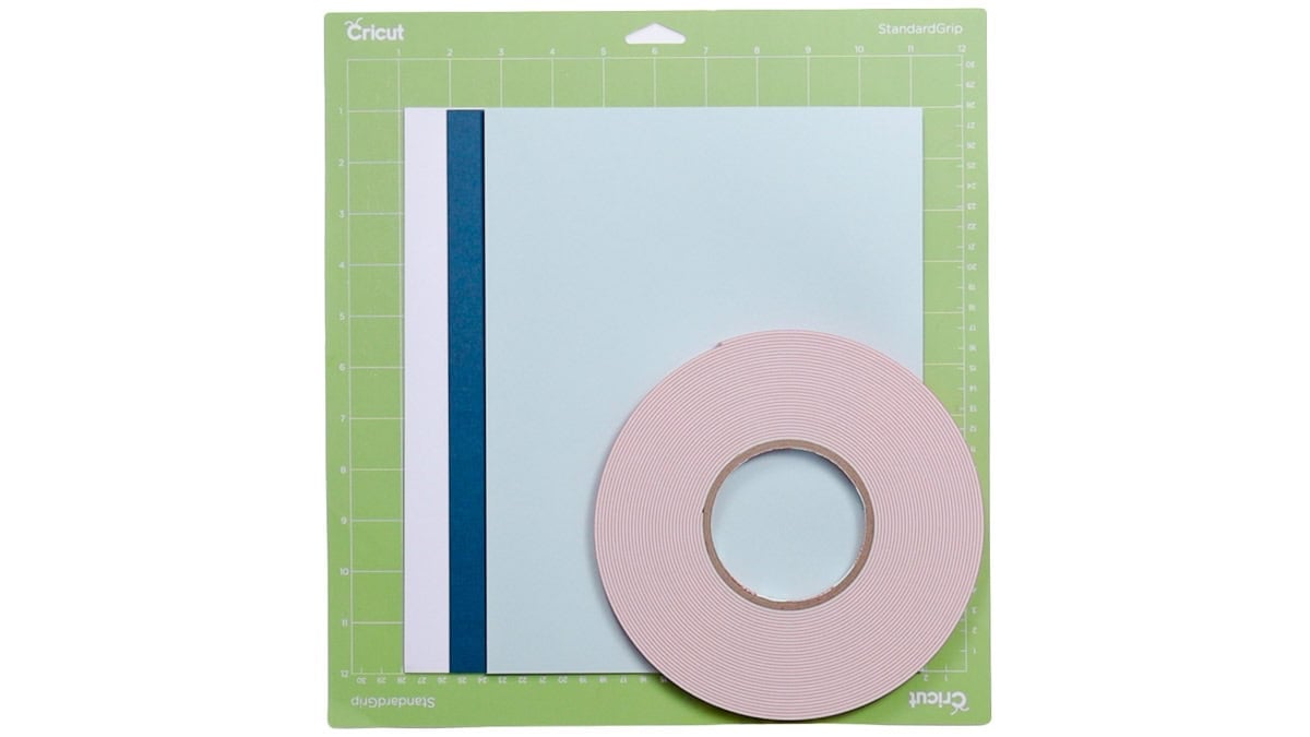 Light blue, dark blue, and white cardstock with a roll of pink foam adhesive tape on a green Cricut StandardGrip cutting mat.