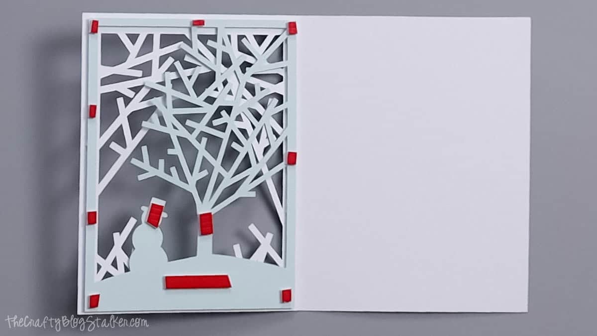 Red foam adhesive strips and squares attached to the back of a layered Christmas card featuring a light blue tree and snowman.