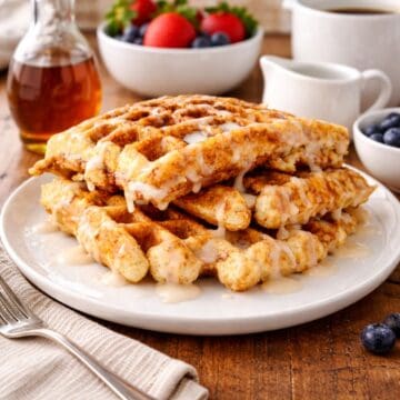 Stack of cinnamon swirl waffles drizzled with icing on a plate, served with syrup, fresh berries, and coffee on a wooden table.