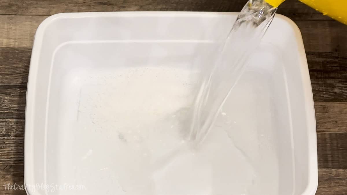 Water being poured from a yellow container into a white plastic tub on a wooden surface to prepare a homemade foot soak.