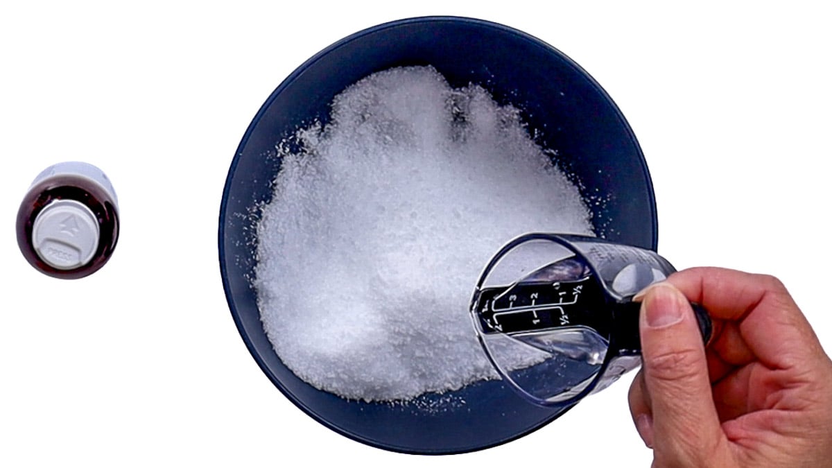 A hand holding a clear measuring cup over a dark blue bowl of Epsom salt while preparing a homemade foot soak with coconut oil.