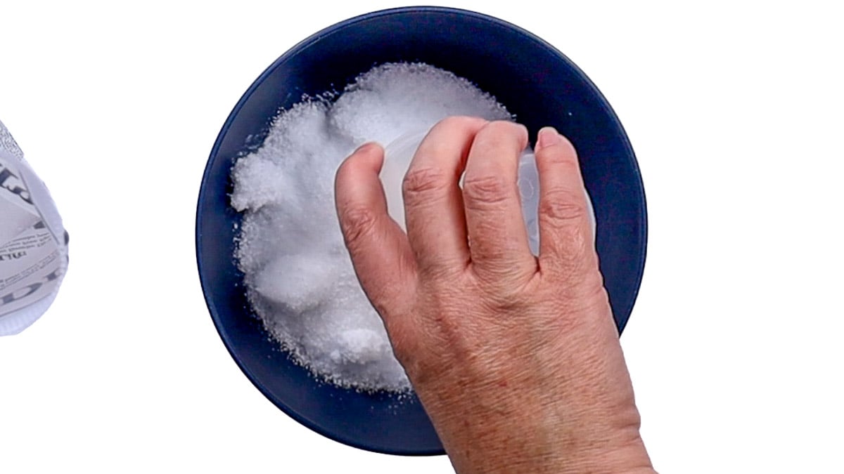 A hand pouring white Epsom salt crystals from a measuring cup into a dark blue bowl to prepare a homemade foot soak.