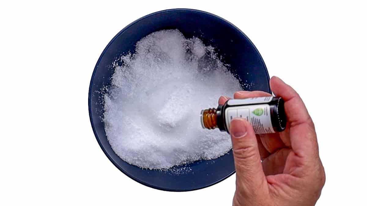 A hand holding an open bottle of essential oil over a dark blue bowl of Epsom salt while preparing a homemade foot soak.
