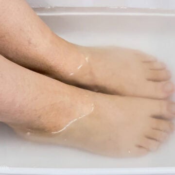 Two bare feet soaking in a white plastic tub filled with cloudy water for a homemade foot soak.