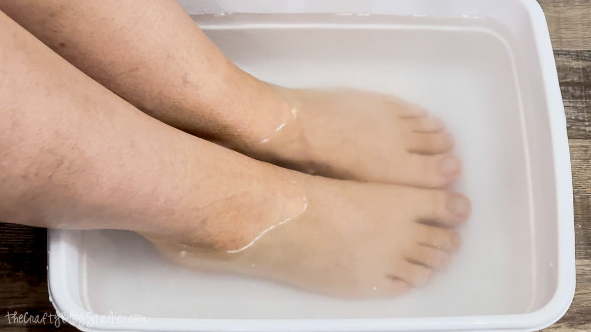Two bare feet soaking in a white plastic tub filled with cloudy water for a homemade foot soak.