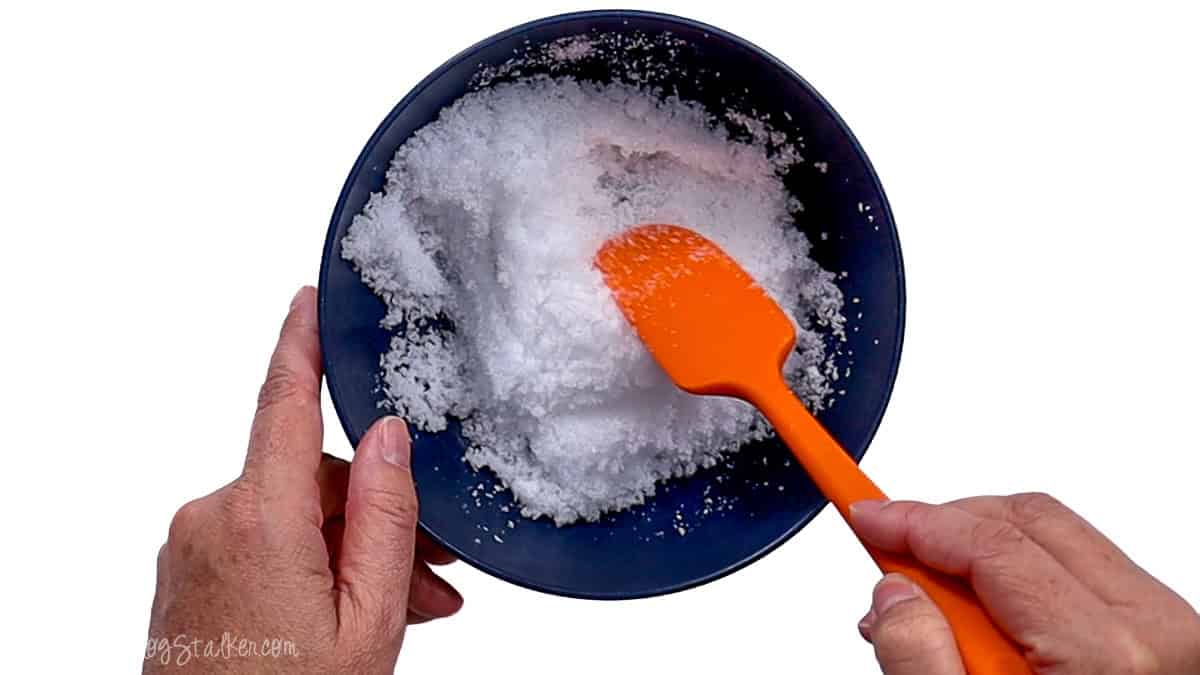 Hands stirring white Epsom salt crystals with an orange spatula in a dark blue bowl while mixing a homemade foot soak.
