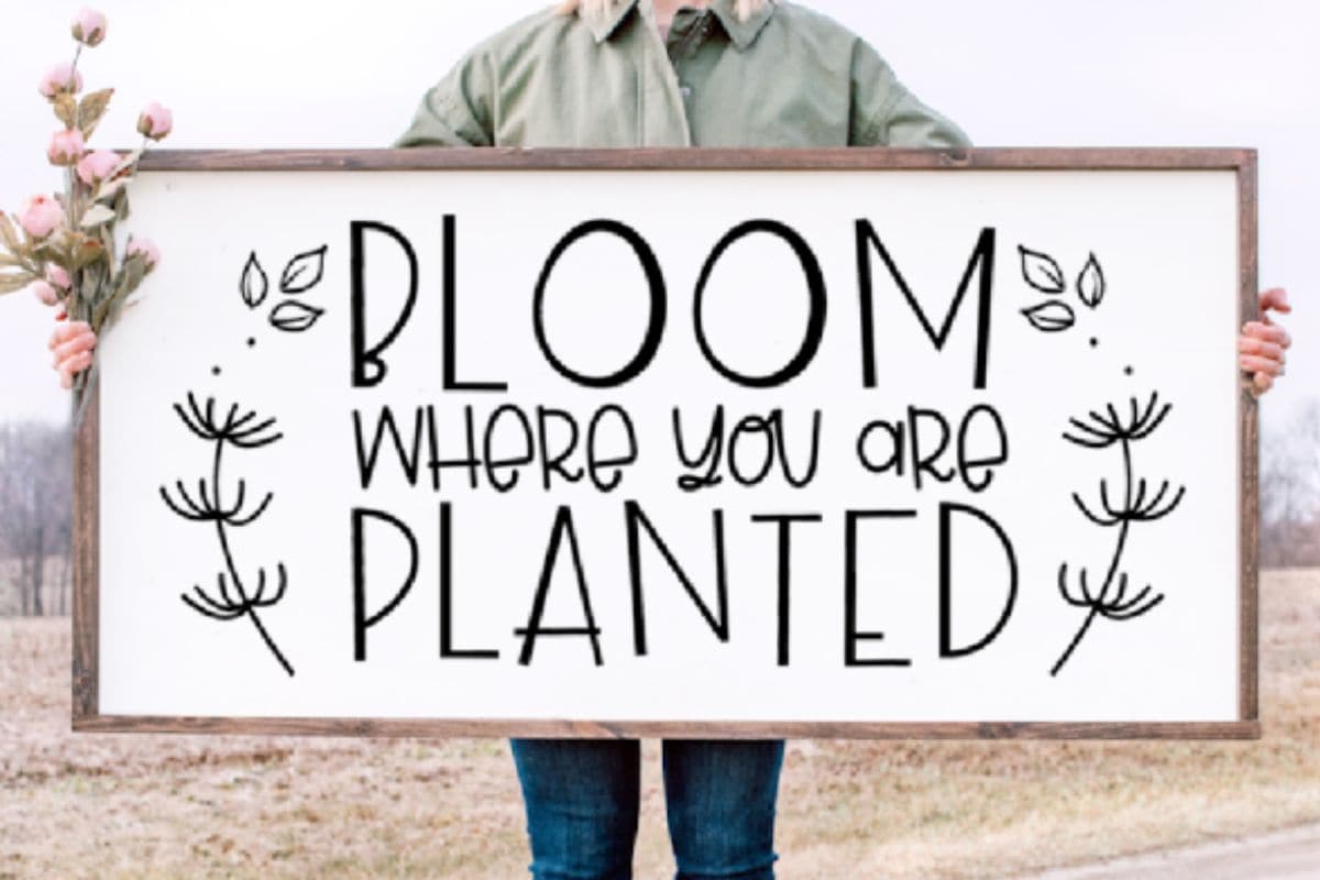 Woman holding a large sign that reads 'bloom where you are planted'.