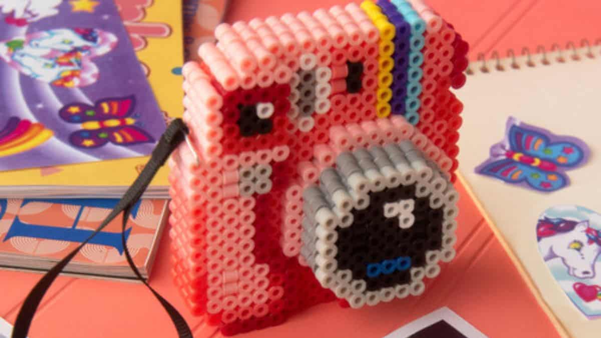 3D instant camera made from colorful Perler beads, featuring pink sides, a rainbow stripe, and a gray lens, displayed on a desk with notebooks and stickers.