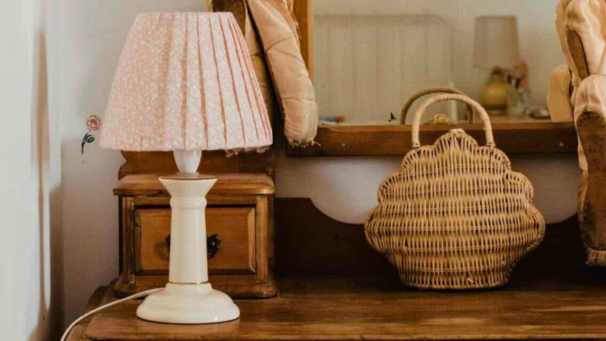 A wooden dresser with a mirror, topped with a white lamp featuring a pleated pink lampshade and a woven wicker handbag beside it.