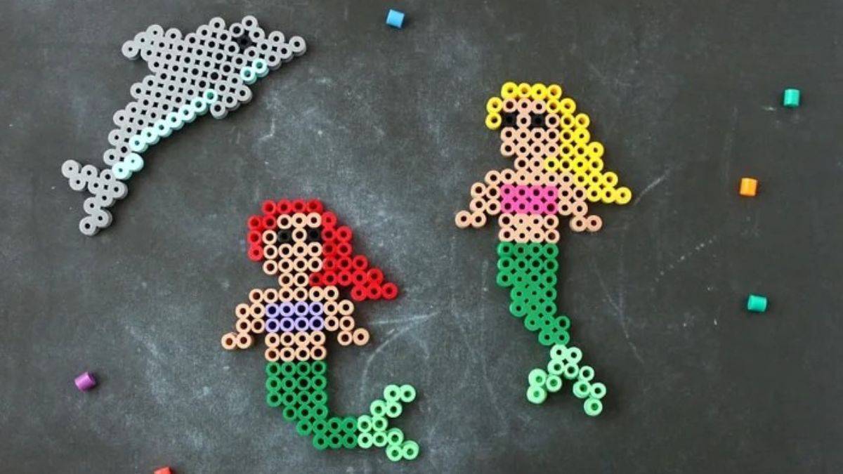 Two mermaids and a dolphin made from colorful Perler beads arranged on a dark surface with scattered loose beads around them.