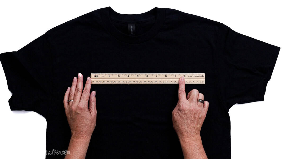 Hands holding a ruler across the chest area of a laid-flat T-shirt to measure placement for a design.