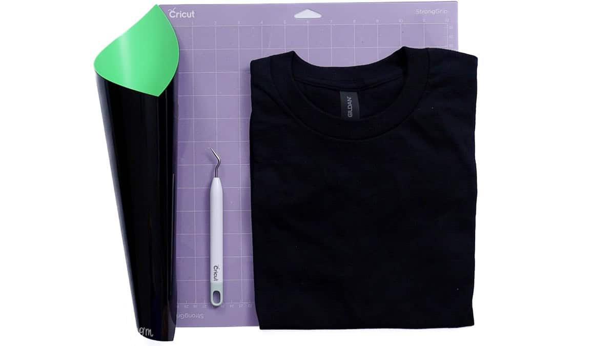 Flatlay of Cricut crafting supplies including a roll of vinyl, a weeding tool, a cutting mat, and a folded black T-shirt.