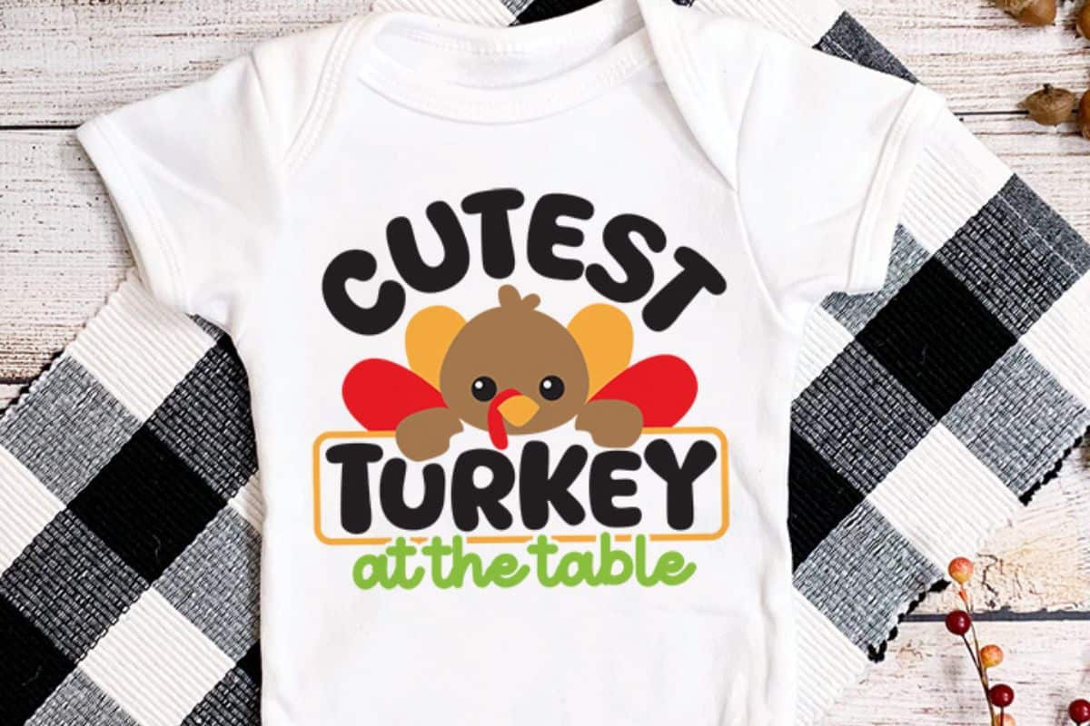 White baby shirt with a turkey design that reads Cutest Turkey At The Table.