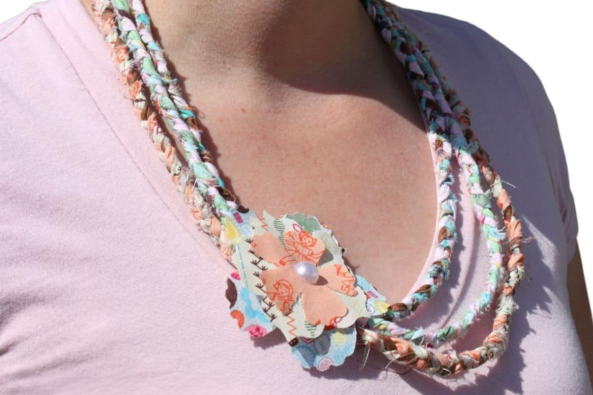 Close-up view of a no-sew braided fabric necklace featuring colorful fabric scraps and a layered fabric flower with a pearl center, adding a handmade touch to a casual outfit.