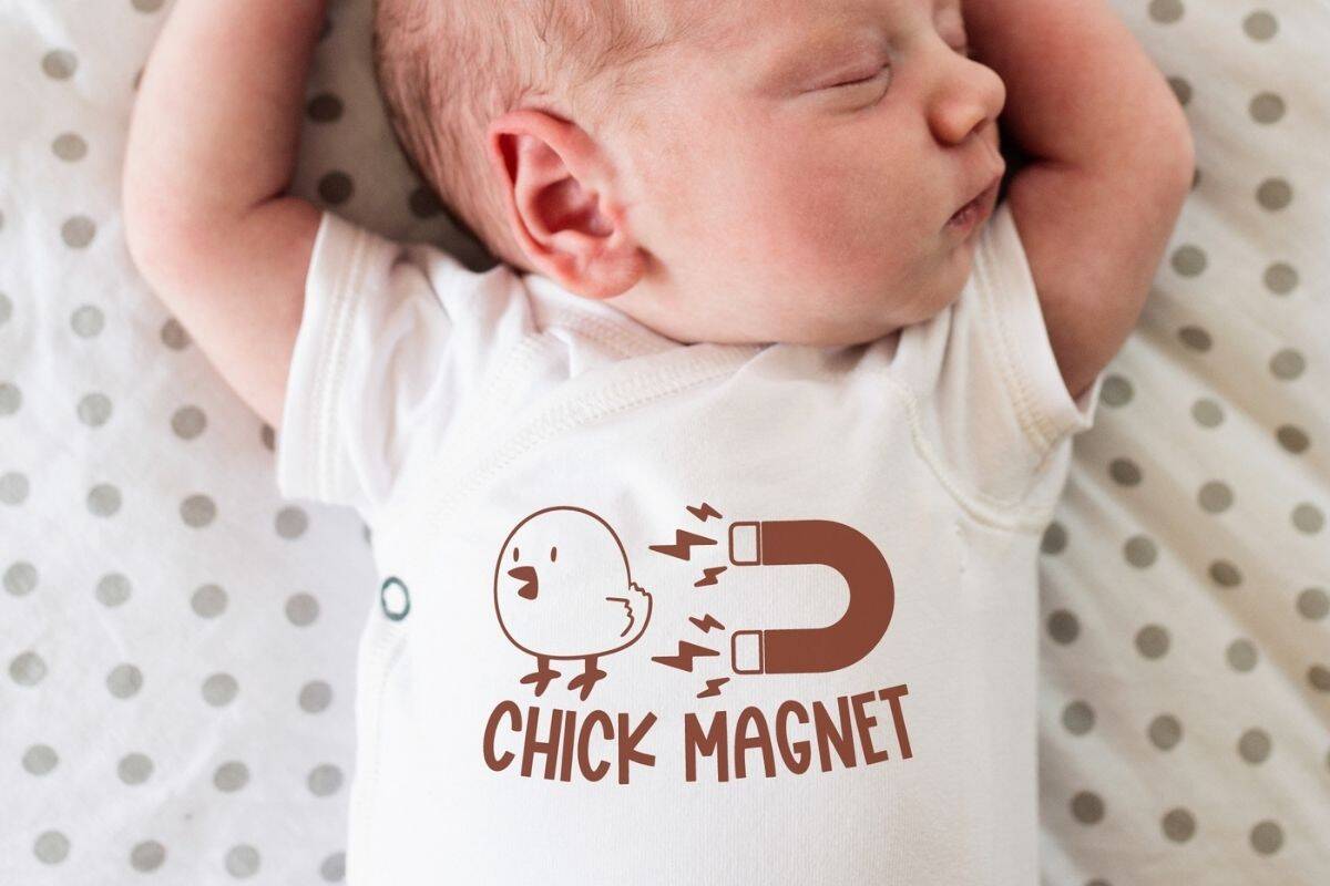 Sleeping baby wearing a white onesie with a cute brown "Chick Magnet" design featuring a chick and a horseshoe magnet with lightning bolts.