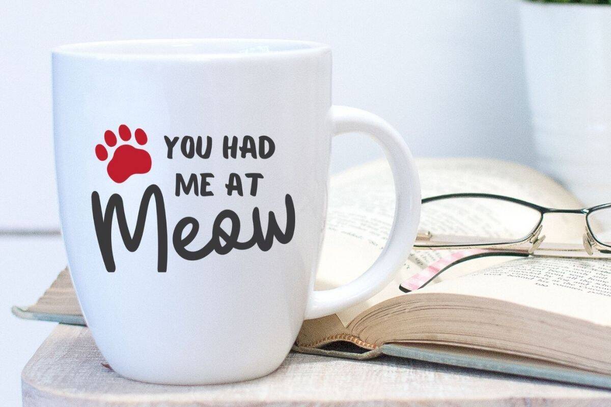 White coffee mug with the phrase “You had me at Meow” in black and a red paw print, placed next to an open book and reading glasses.