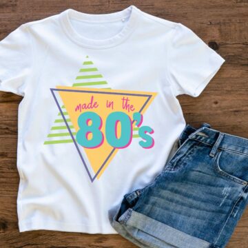 White t-shirt with a retro "Made in the 80’s" design featuring colorful geometric shapes, paired with denim shorts on a wooden background.