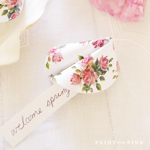 Floral paper fortune cookie with 'Welcome Spring' message, placed on a white tablecloth with pink roses and lisianthus flowers in the background.
