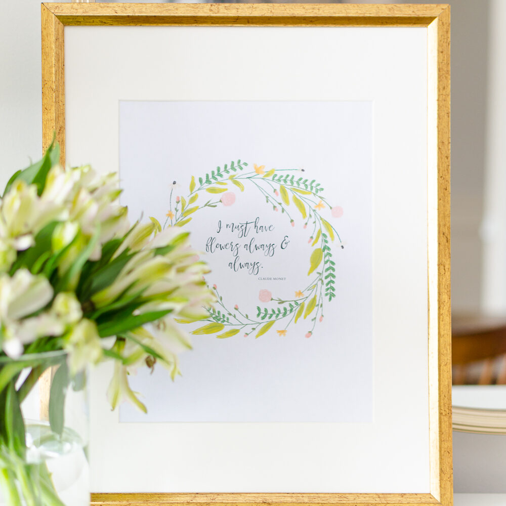 Framed spring printable with a floral wreath and Claude Monet quote, placed on a white surface with a vase of fresh white flowers in the foreground.