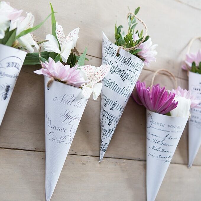 Vintage-style paper cones with printed sheet music and script, filled with fresh pink and white flowers, arranged on a rustic wooden surface.