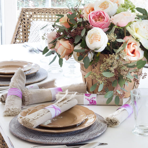 Elegant spring table setting with a floral centerpiece in a wooden bucket, neutral dishware, frayed linen napkins, and decorative napkin rings with pink accents.