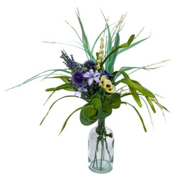 Artificial flower bouquet in a clear glass vase with purple, cream, and green stems arranged with greenery and wispy grass against a white background.